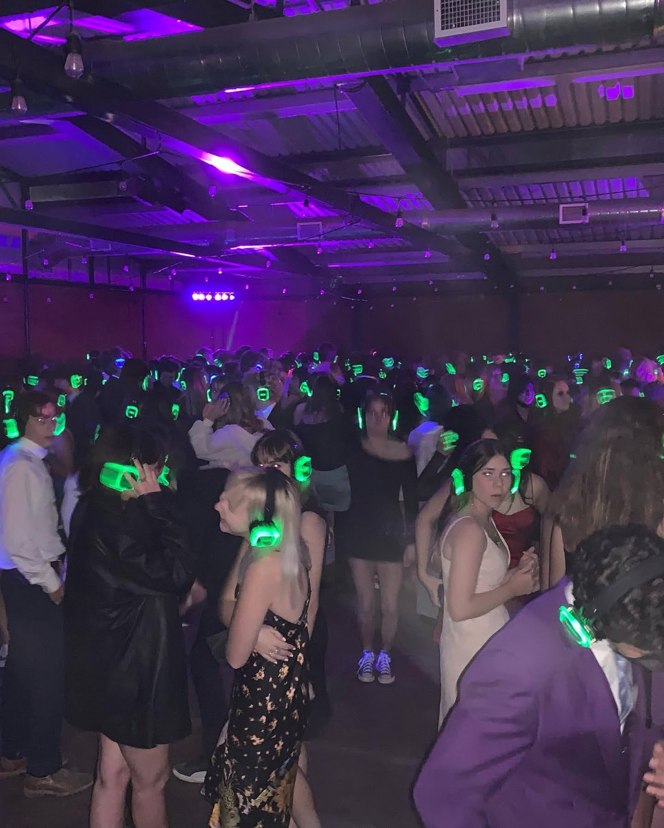 A large group of people are dancing in a dimly lit room, all wearing glowing green headphones for a silent disco. Purple lighting adds atmosphere, reminiscent of the vibrant colors at the Flower Fields Carlsbad, and the crowd is dressed in semi-formal attire.