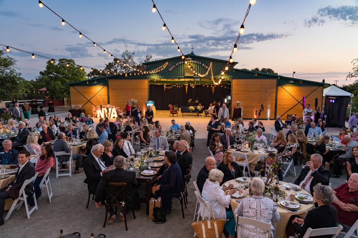 A large outdoor evening event at the flower fields Carlsbad, with many people seated at round tables under string lights, enjoying dinner in front of a barn adorned with lights and large illuminated NVA letters.