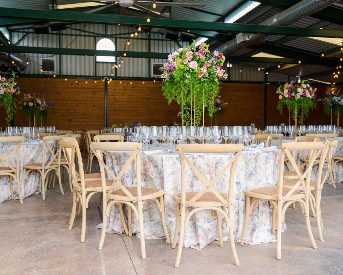 Round tables with floral tablecloths are set for an event, each decorated with tall green and pink floral centerpieces reminiscent of The Flower Fields Carlsbad. Wooden chairs surround the tables in a spacious hall with string lights and a wooden wall.