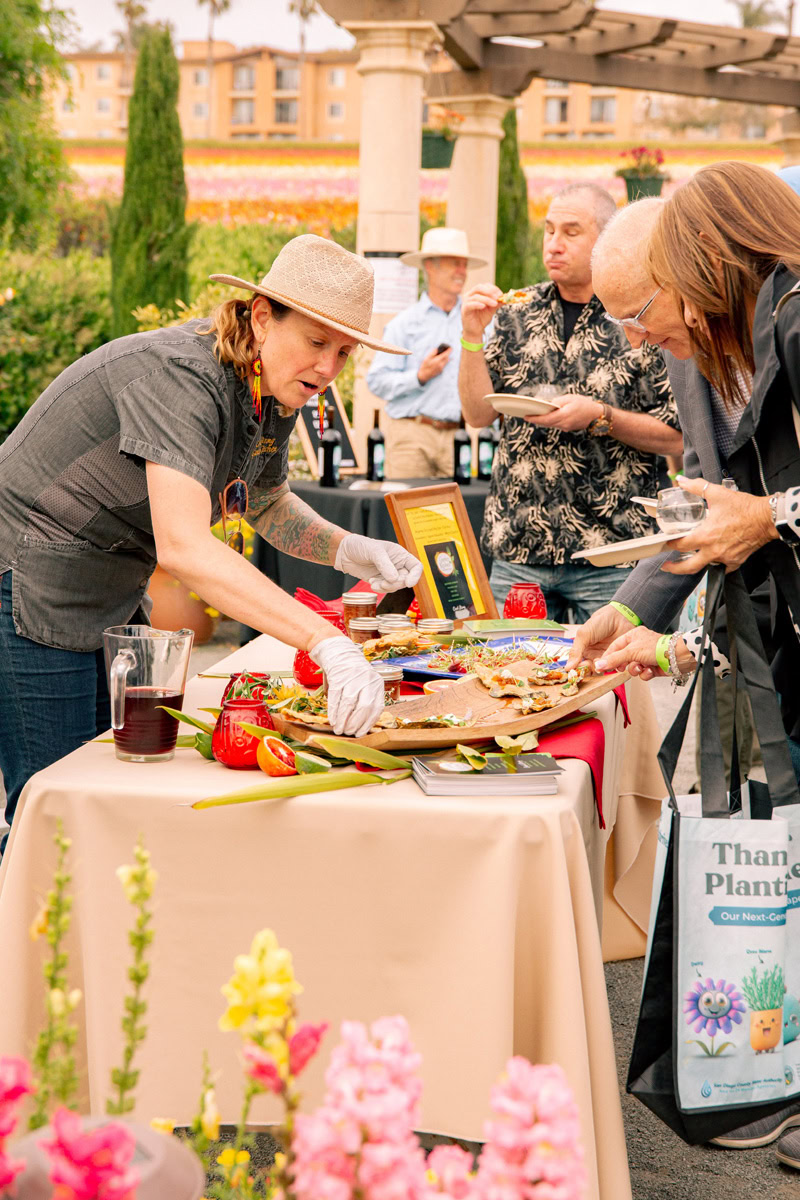 A woman in a hat and gloves serves food samples to event attendees at an outdoor table decorated with vegetables at The Flower Fields Carlsbad. People mingle, holding plates, surrounded by vibrant flowers and lush greenery in the background.