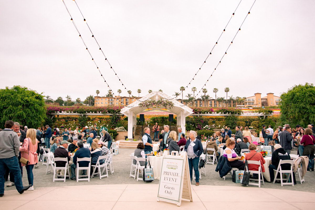 An outdoor event at The Flower Fields Carlsbad, with guests seated at round tables under string lights near a white gazebo. A sign in front reads &ldquo;Private Event, Guests Only.&rdquo; Buildings and palm trees can be seen in the background.