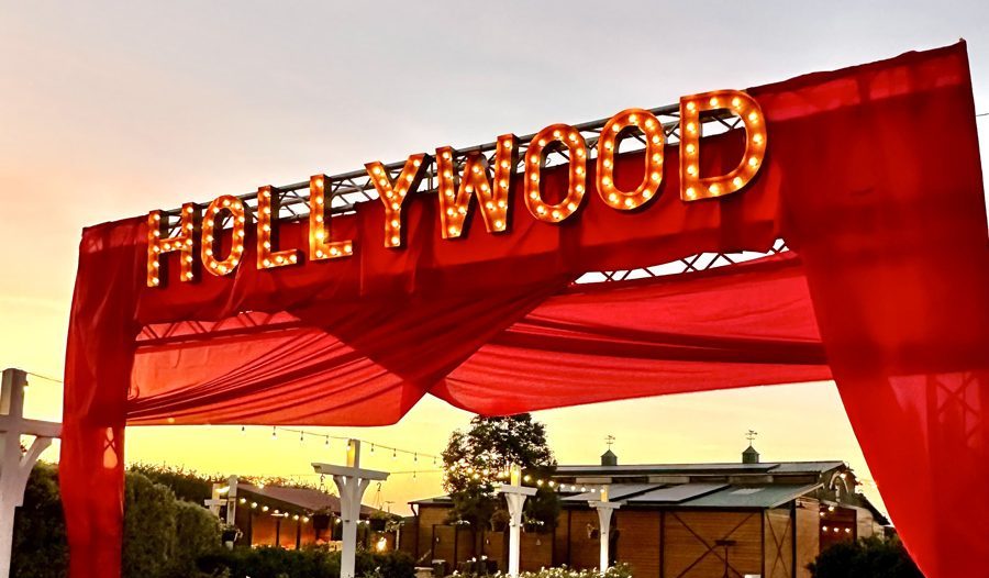 A large red entrance at The Flower Fields Carlsbad, decorated with lights spelling HOLLYWOOD, stands over a red carpet and velvet rope, creating a glamorous, movie-premiere-style walkway outdoors at dusk.