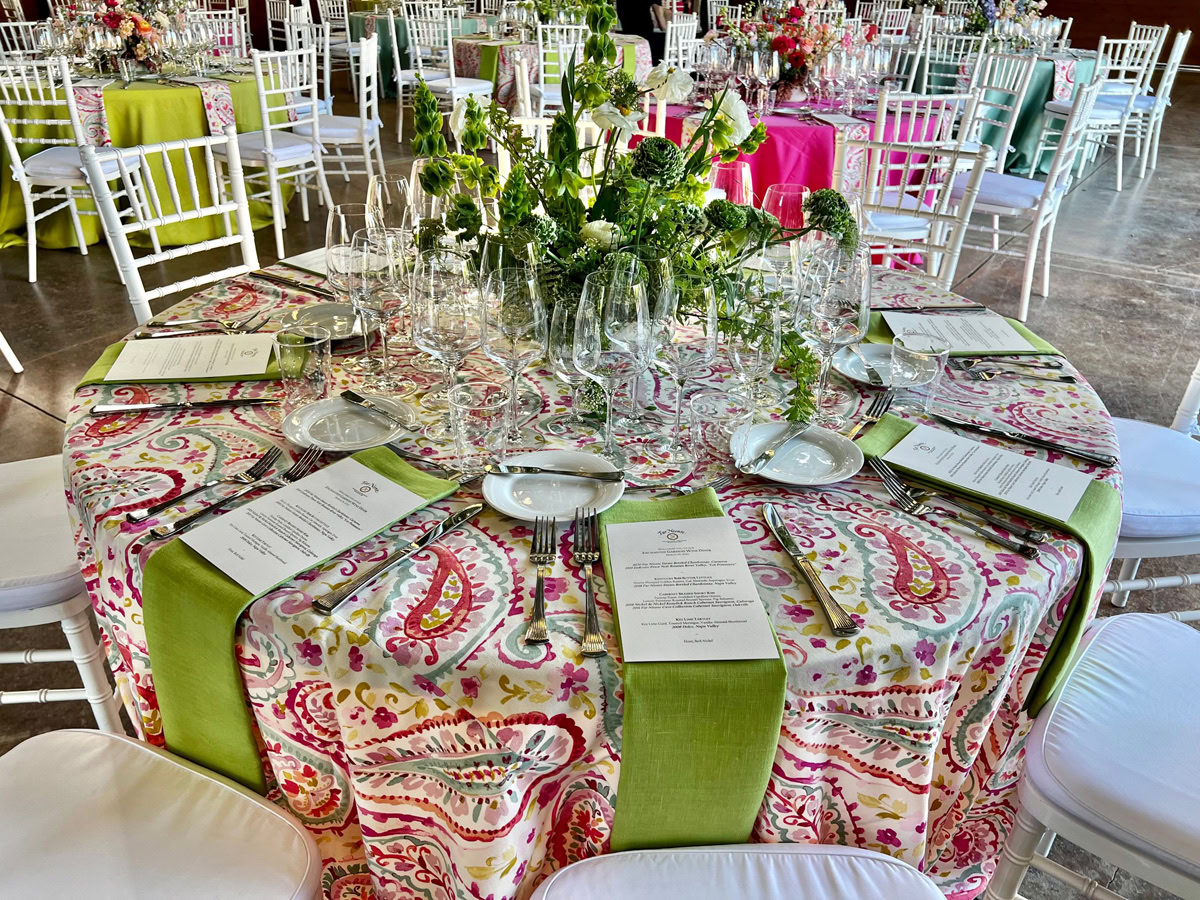 A round table set for a formal event, featuring a colorful paisley tablecloth reminiscent of The Flower Fields Carlsbad, a green table runner, menus at each place setting, glassware, and a centerpiece of green foliage, surrounded by white chairs.