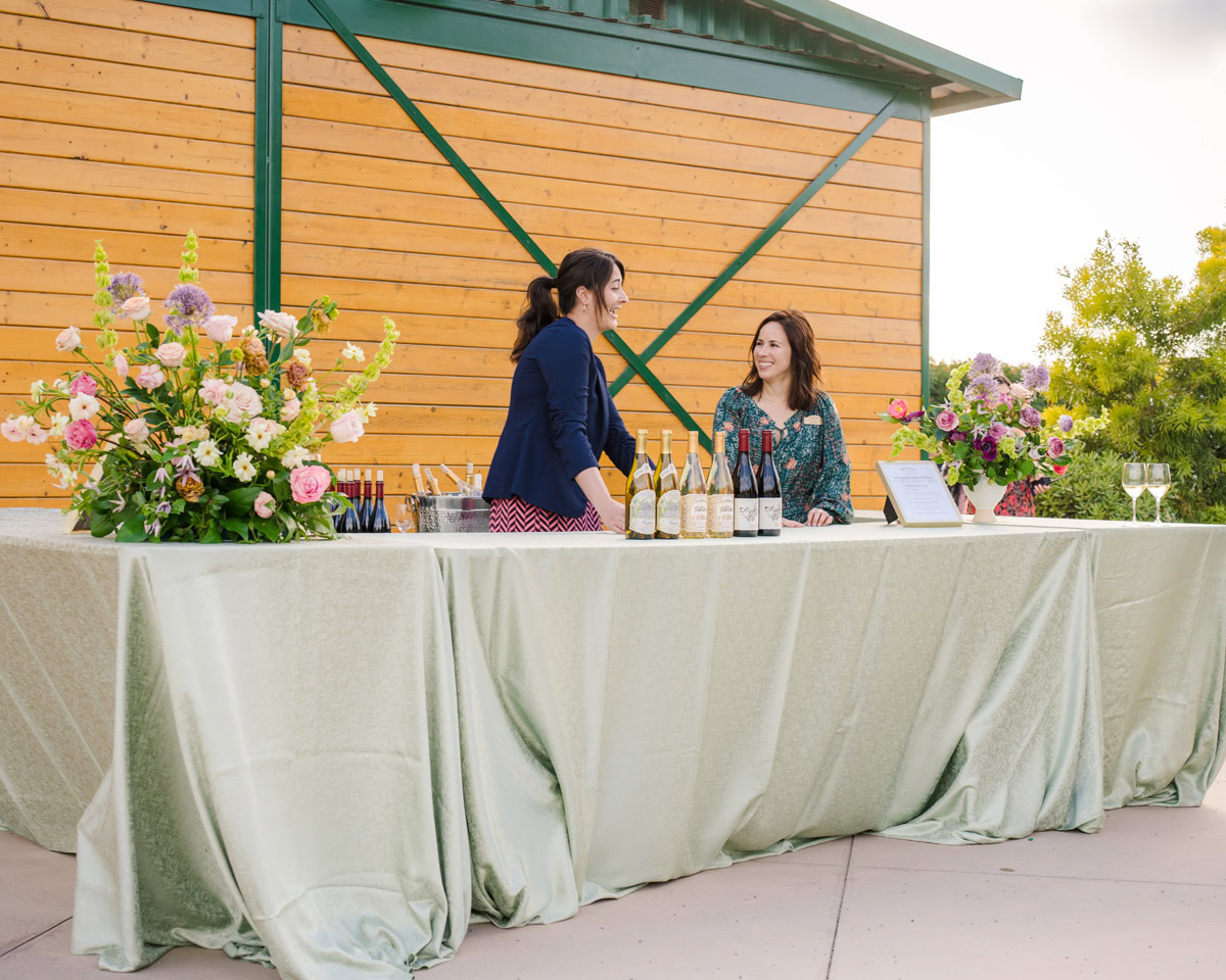 Two women stand behind a long table covered with a light green tablecloth, displaying wine bottles and floral arrangements, smiling and chatting in front of a wooden building at The Flower Fields Carlsbad.
