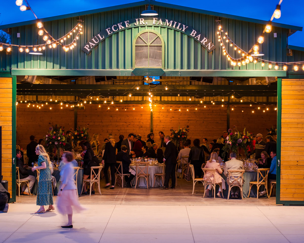 Guests gather at decorated tables inside the barn venue at The Flower Fields Carlsbad, with string lights overhead at dusk. The sign reads Paul Ecke Jr. Family Barn. A few people walk outside the entrance, adding to the festive and warm atmosphere.