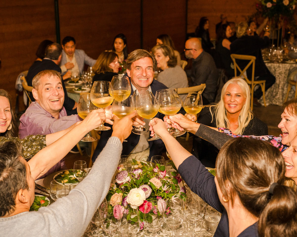 A group of people sitting around a table at a festive indoor gathering clink their glasses in a toast, smiling and surrounded by flowers and dinnerware inspired by the vibrant blooms of The Flower Fields Carlsbad.