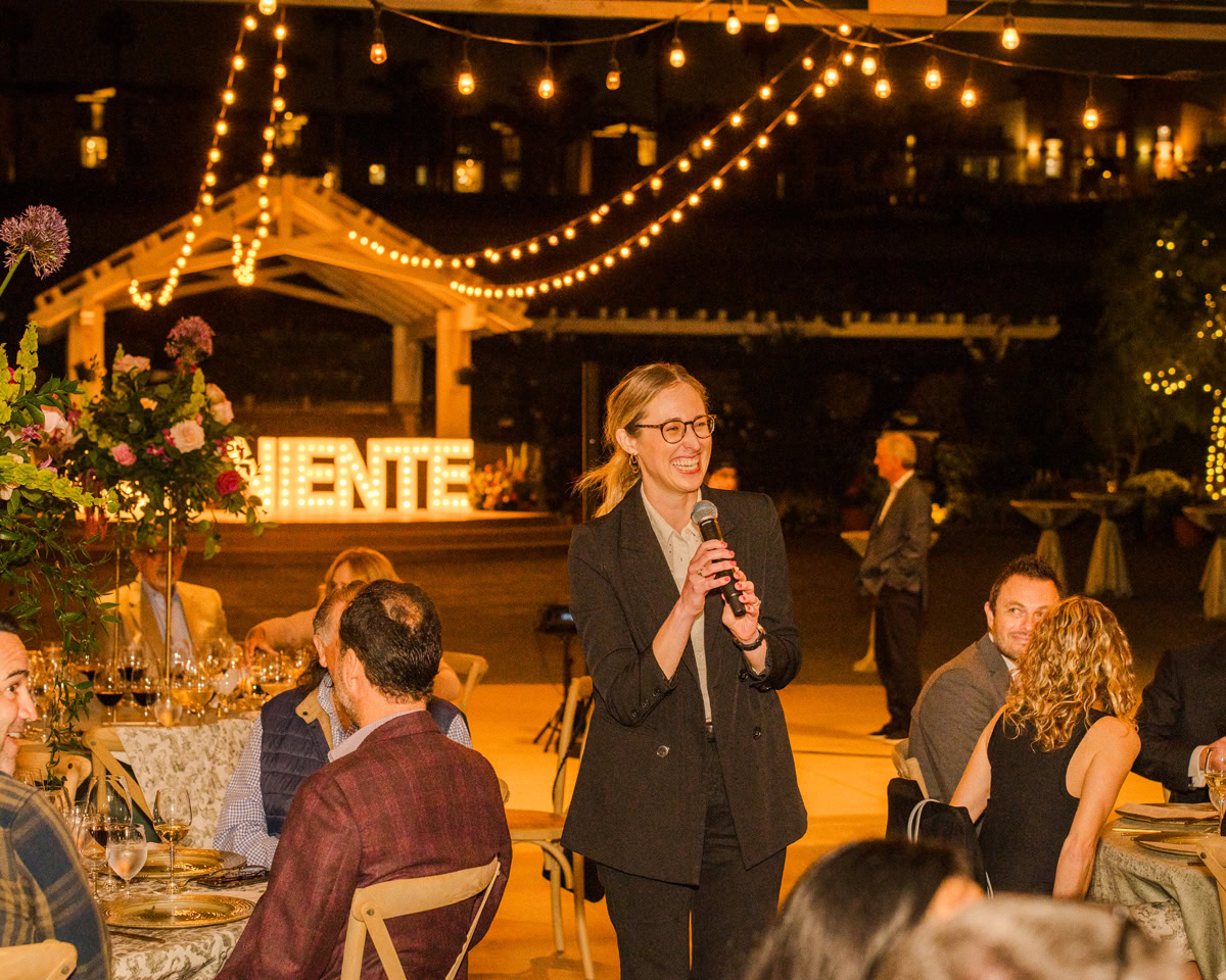 A woman in a suit speaks into a microphone at an outdoor evening event at the Flower Fields Carlsbad, with string lights overhead. Guests seated at tables listen as illuminated letters spelling ENTE shine in the background.