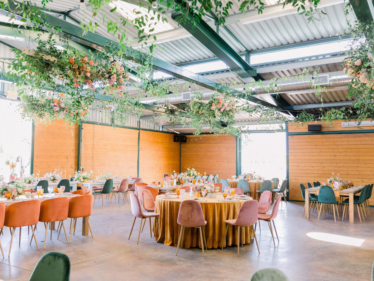 A bright, airy event space with wooden walls, decorated with hanging greenery and flowers inspired by the Flower Fields Carlsbad. Round tables gleam with gold tablecloths, surrounded by pink and green velvet chairs, each topped with floral centerpieces.
