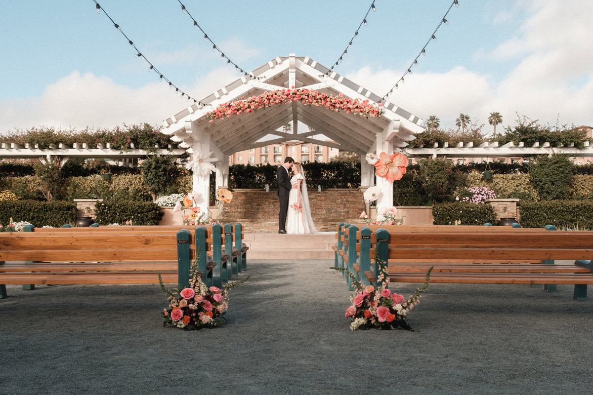 Bride and groom standing under a decorated white pergola with floral arrangements at The Flower Fields Carlsbad.