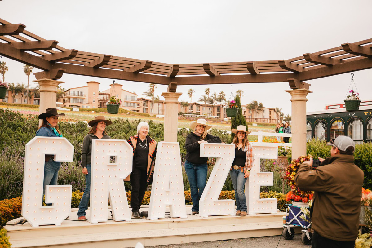 Five people wearing cowboy hats pose in front of large marquee letters spelling GRAZE under a pergola outdoors at The Flower Fields Carlsbad, while another person snaps their photo. Residential buildings and lush greenery frame the scene.