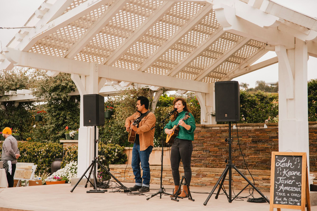 Two musicians perform on an outdoor stage under a white pergola at The Flower Fields Carlsbad. The man plays guitar while the woman sings and plays ukulele, with a chalkboard sign displaying their names amid colorful shrubs and blooms in the background.