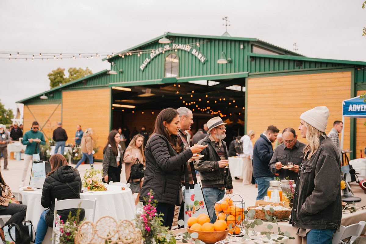 People enjoy food and drinks at outdoor tables in front of a green barn during a lively market or festival at The Flower Fields Carlsbad. Flowers, oranges, and strings of lights create a festive atmosphere.
