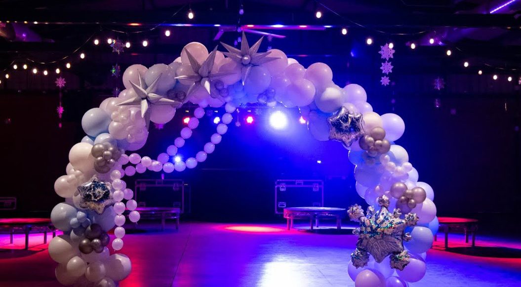 A balloon arch with white and silver decorations stands under string lights in front of the Paul Ecke Jr. Family Barn entrance at the Flower Fields Carlsbad, illuminated by colorful lights.