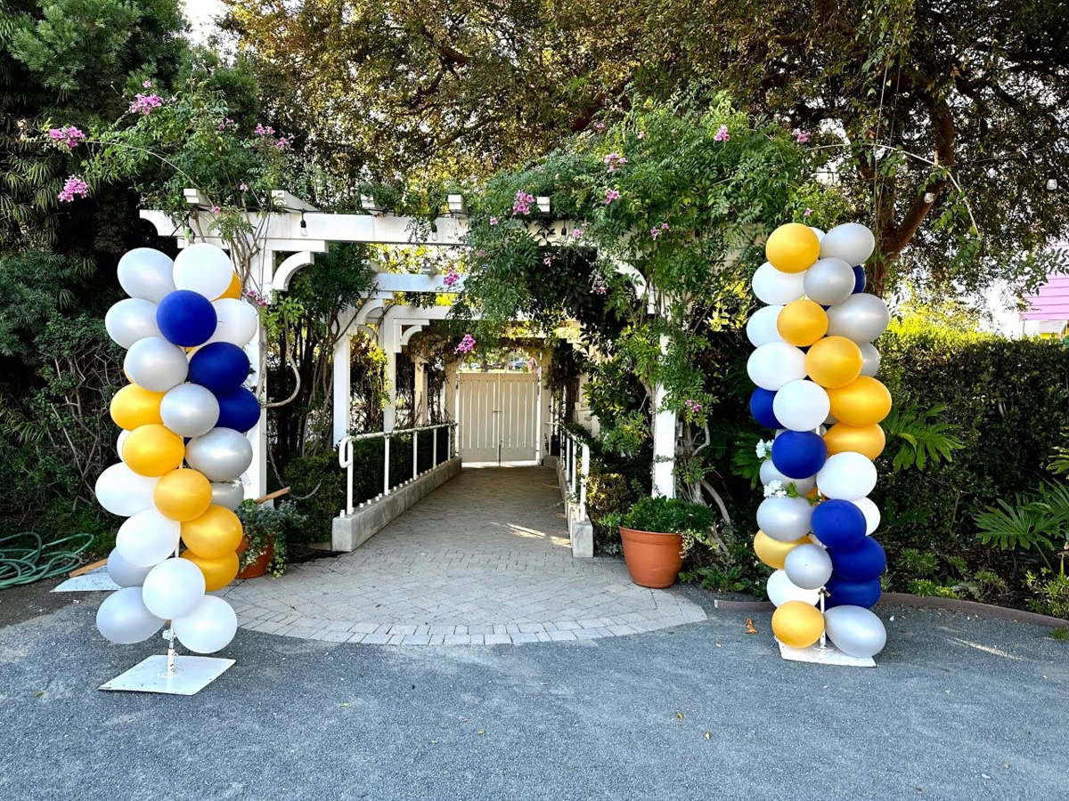 A white pergola decorated with flowers and greenery stands at the end of a paved path, flanked by columns of blue, gold, silver, and white balloons beneath leafy trees, evoking the vibrant beauty of The Flower Fields Carlsbad.