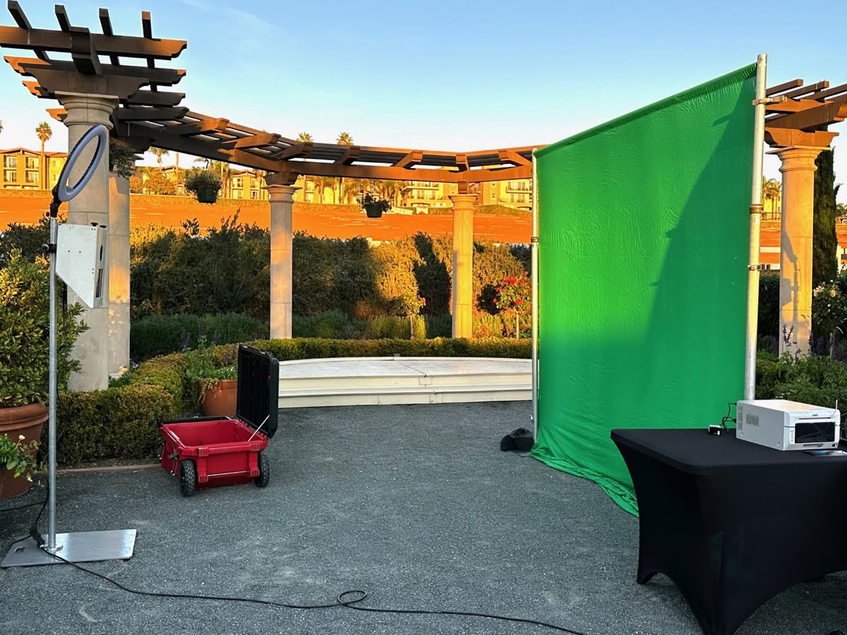 A green screen stands outdoors next to a black table with a white device, a red wagon, and a circular sign at The Flower Fields Carlsbad, with pergolas, plants, and buildings in the background under evening sunlight.