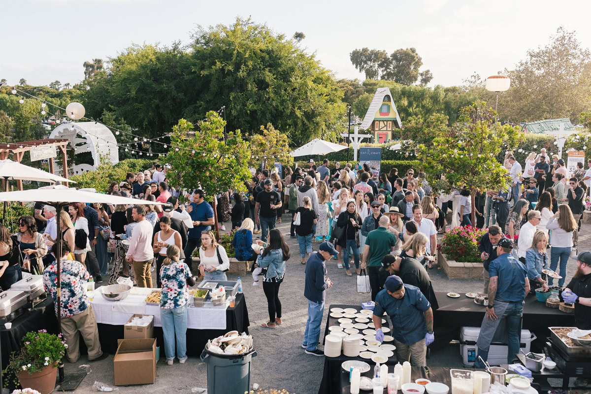 A large crowd gathers outdoors at a lively event with food stations, tables, and string lights. People are mingling and serving food, enjoying the sunny weather surrounded by trees, greenery, and the vibrant blooms of the Flower Fields Carlsbad.